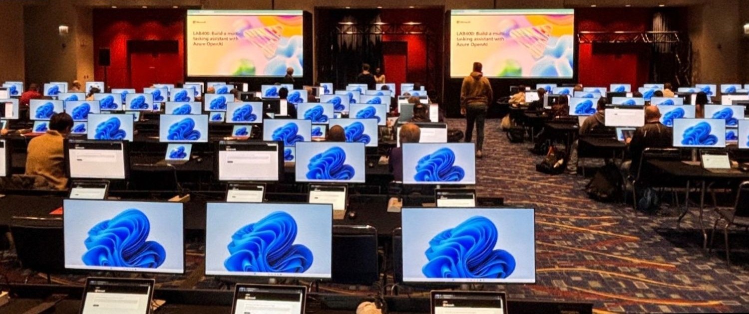Ein Konferenzraum mit sehr vielen Arbeitsplätzen; einige Menschen sitzen an den Arbeitsplätzen und schauen auf die Monitore